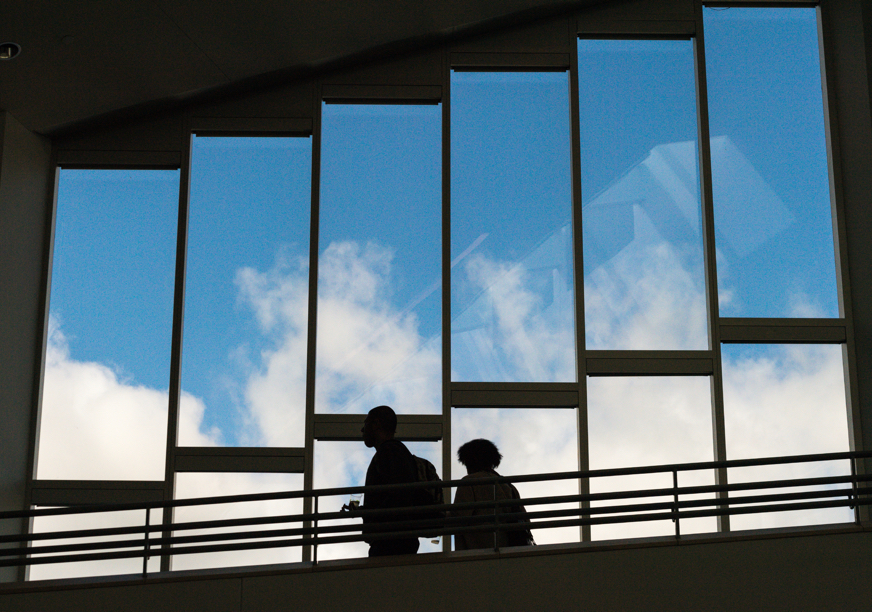 In a campus building, two students walk past windows that show a blue sky outside.