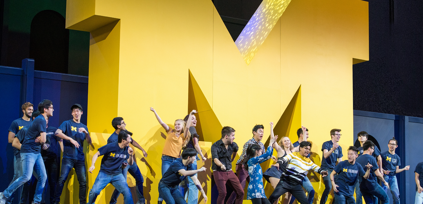 U-M students dance onstage in front of a big yellow Block M.