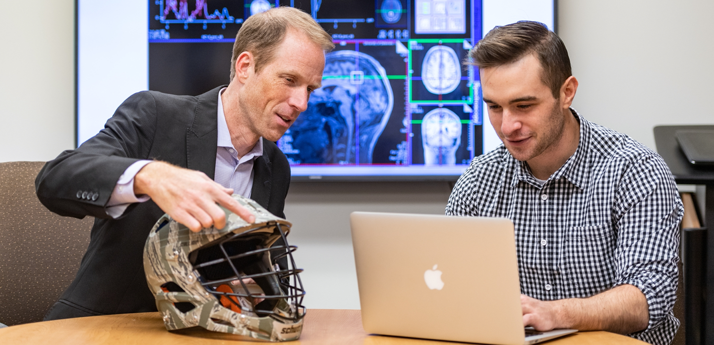 Two U-M researchers examine a football helmet, with a slide of concussion scans in the background.