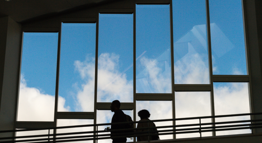 In a campus building, two students walk past windows that show a blue sky outside.